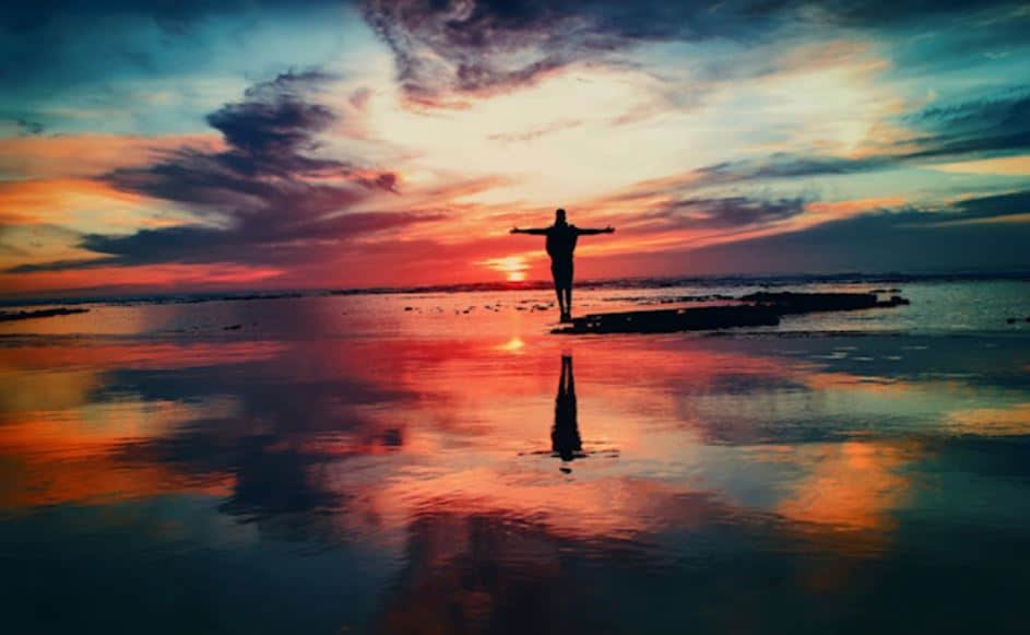 Man standing with arms stretched out on the beach with a brightly colored sunset.