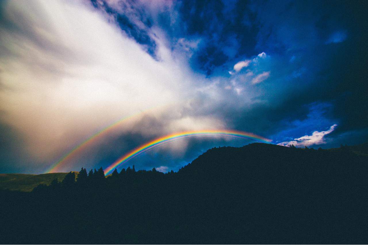 The sun shining through blue clouds over a hill with two rainbows