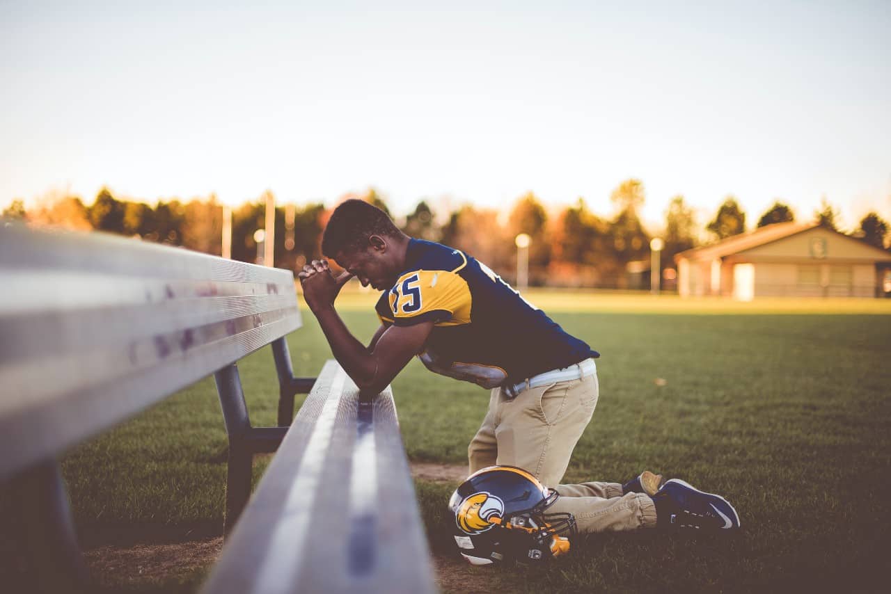 A football player kneeling in prayer on a bench on a football field.