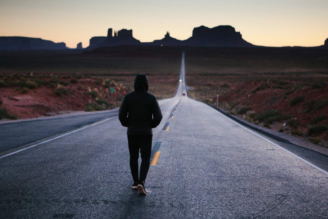 A man in black hoodie walking down a road with rock formations on the horizon at sunset.