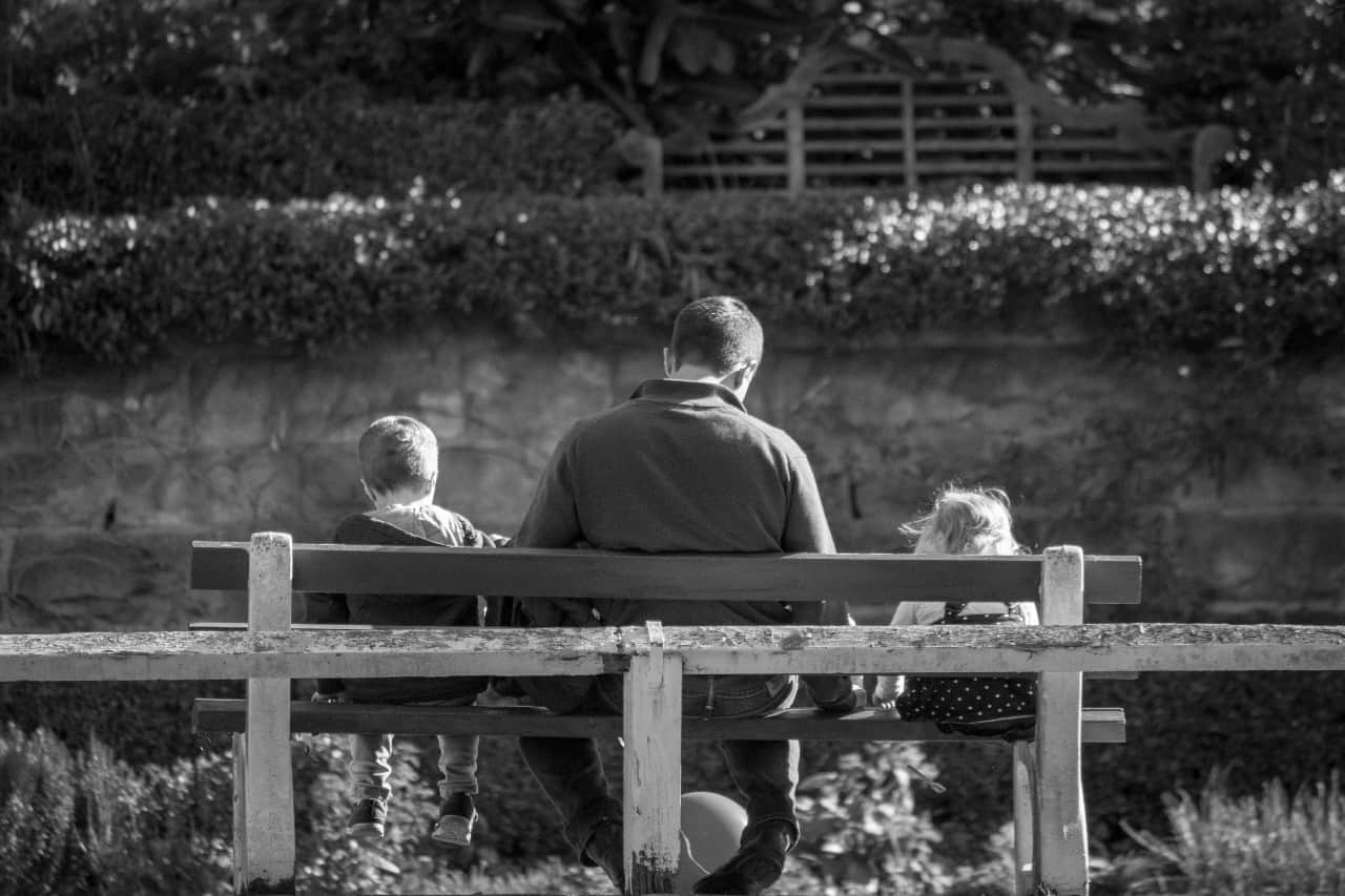 Black and white photo of dad, daughter, and son sitting on a bench with their backs facing the camera.