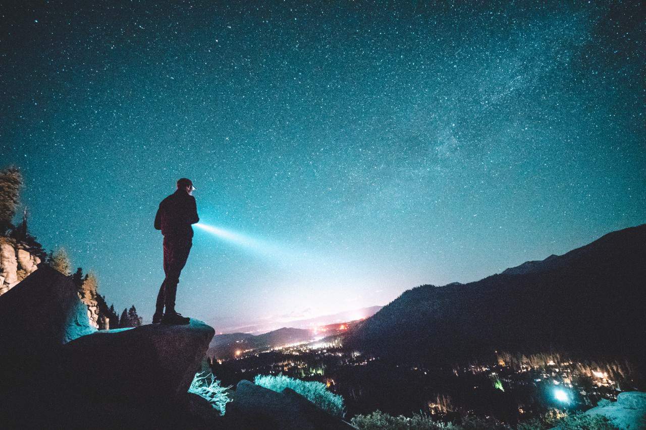 A man searching with a flashlight under a night sky in the mountains.