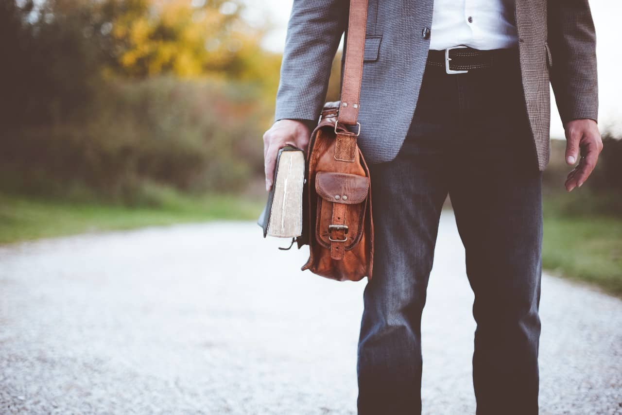 A man with bag on his shoulder and a Bible in his hand walking down a road.