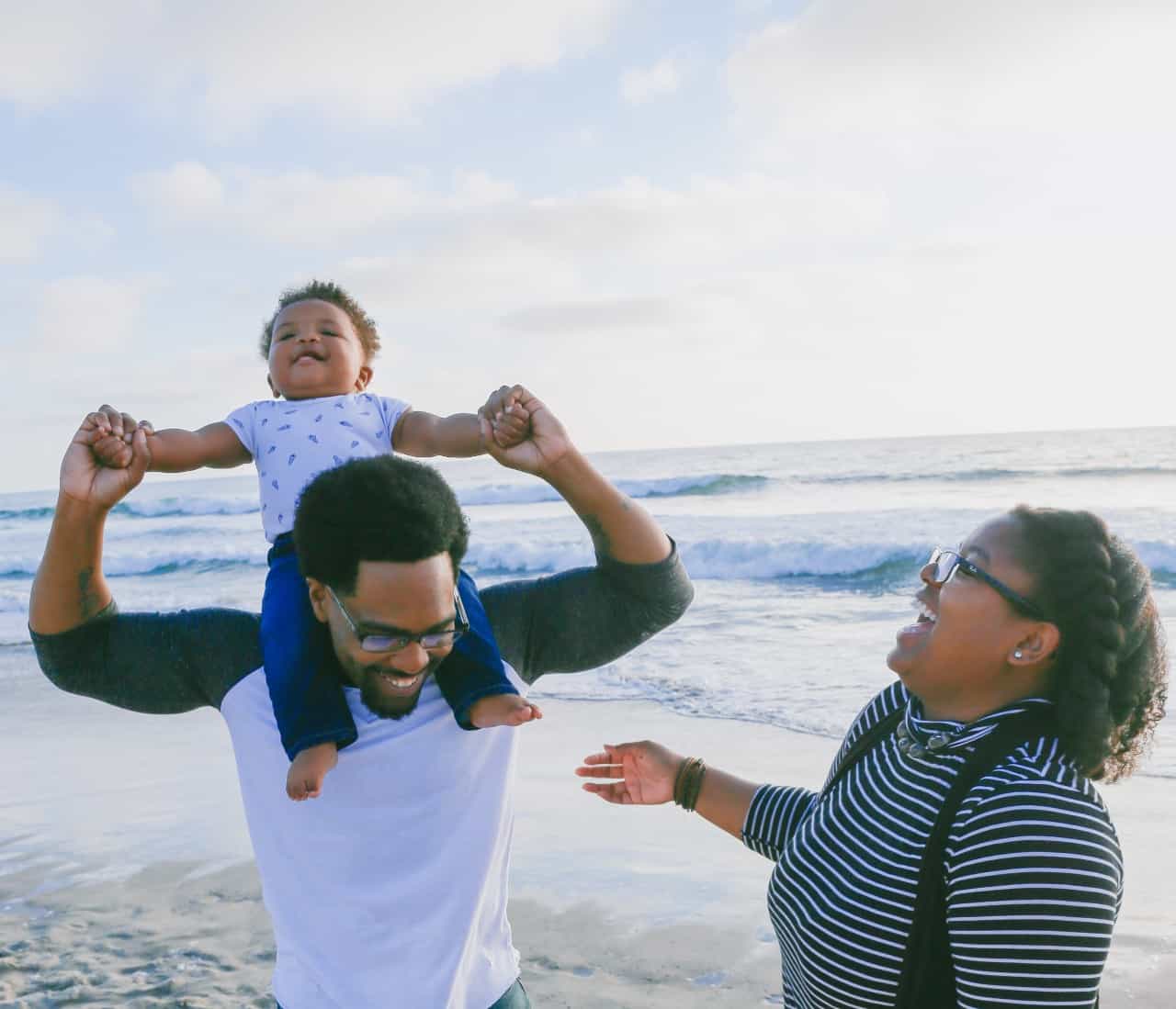 A dad with his baby son sitting on his shoulders and a Mom laughing while they stand on a beach.