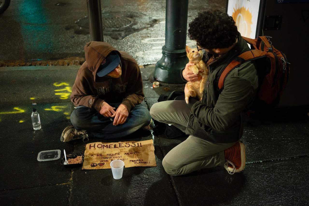 A man kneeling down next to a homeless man and holding the homeless man's cat.