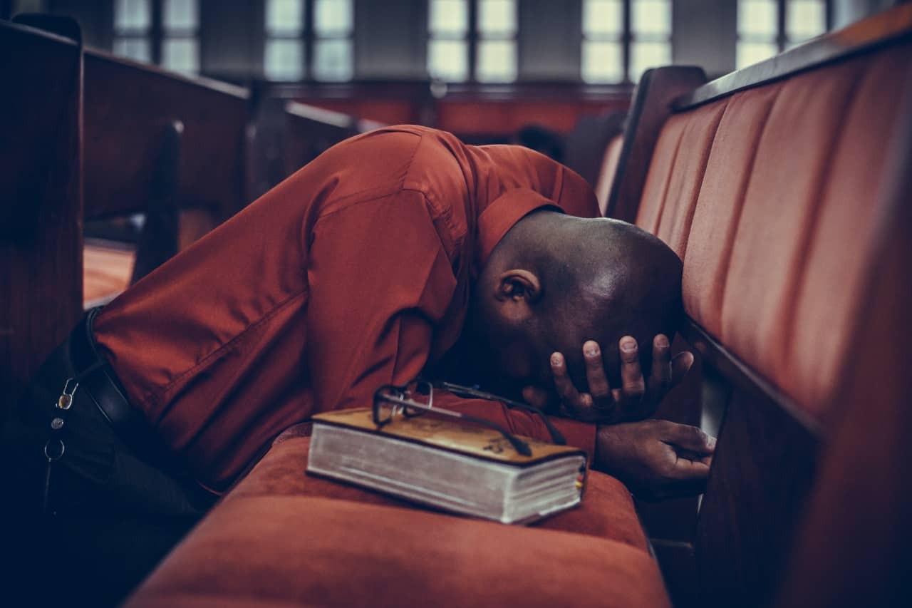 A man on his knees, holding his head, over a church pew with a Bible next to him.