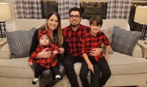 Family picture sitting on couch wearing red and black plaid. mom, dad, 1 boy, and 1 girl.