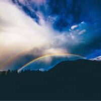 The sun shining through blue clouds over a hill with two rainbows