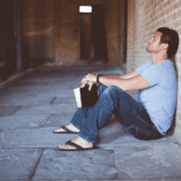 A man sitting on the ground with his back on the wall holding a Bible.