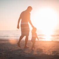 A father and his young daughter holding hands on the beach waking towards the water at sunset.