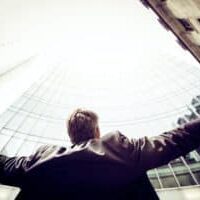 A man in a suit looking up at a skyscraper with arms open wide.