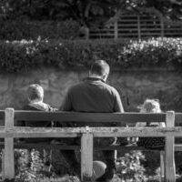 Black and white photo of dad, daughter, and son sitting on a bench with their backs facing the camera.