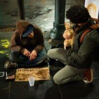 A man kneeling down next to a homeless man and holding the homeless man's cat.